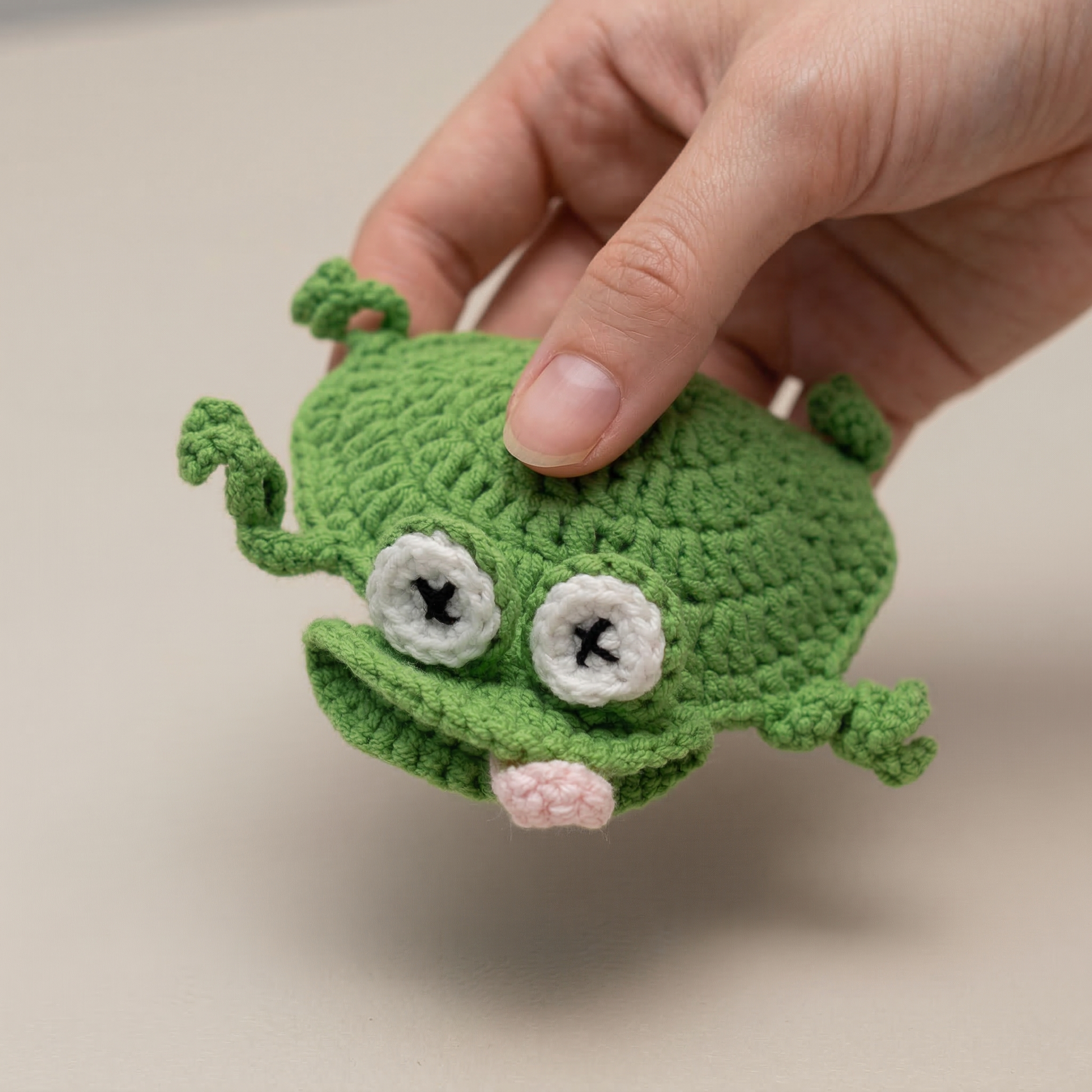 Hand holding a green crocheted frog toy on a beige background