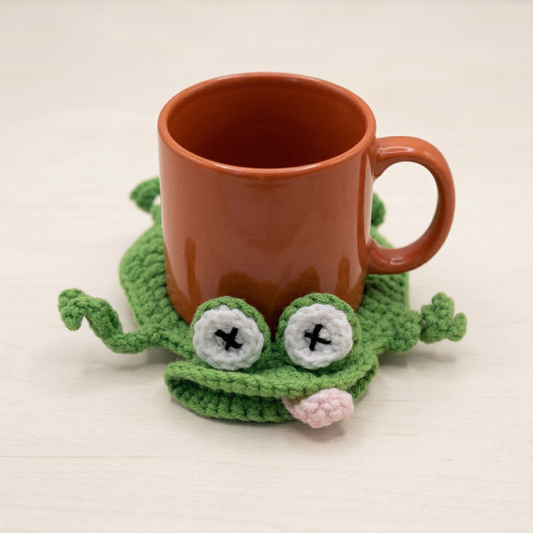 Brown mug on a green frog-shaped crocheted coaster with a white background