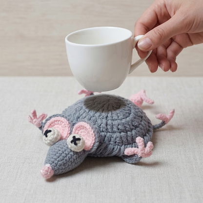 Handmade crochet mouse with pink accents next to a white mug on a light surface.